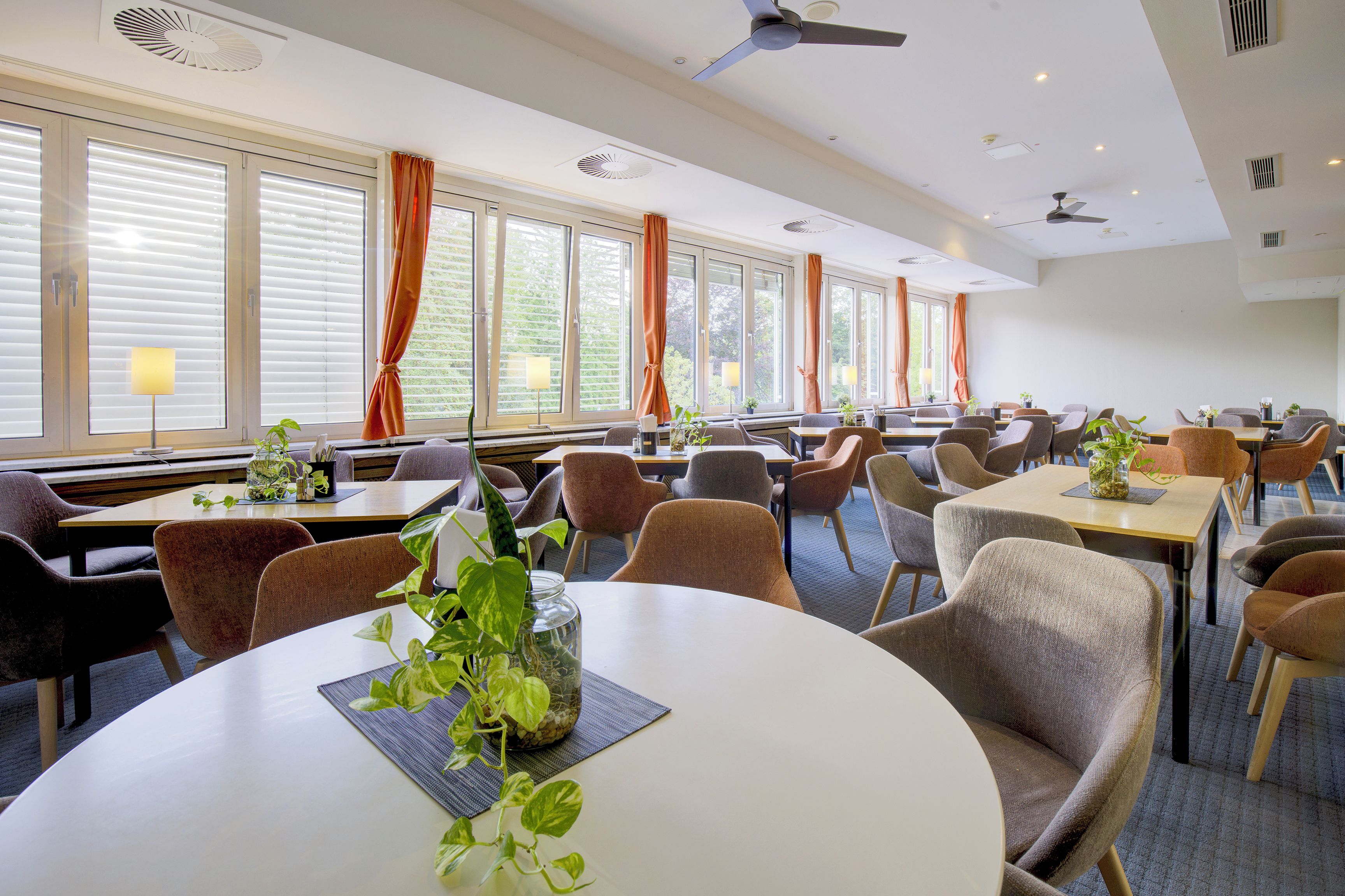 Un comedor de hotel con mesas redondas, sillas y ventanas amplias con cortinas.