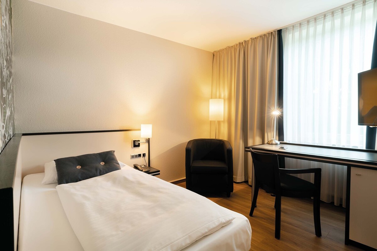 A hotel room with a bed, a desk, a chair, a lamp, and a window with curtains.