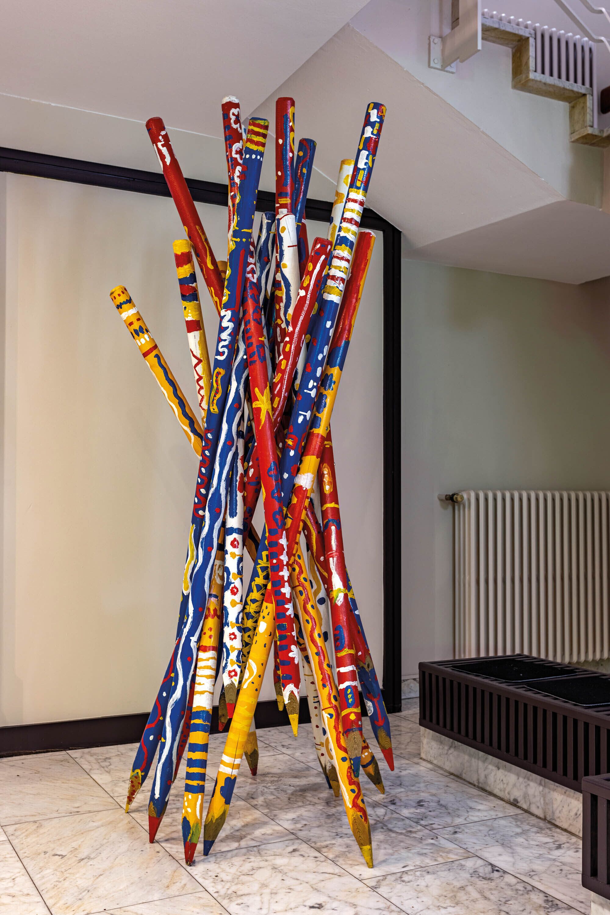 Una scultura di pali dipinti in modo astratto e colorato in un’area hall con pavimento piastrellato e termosifone sullo sfondo.