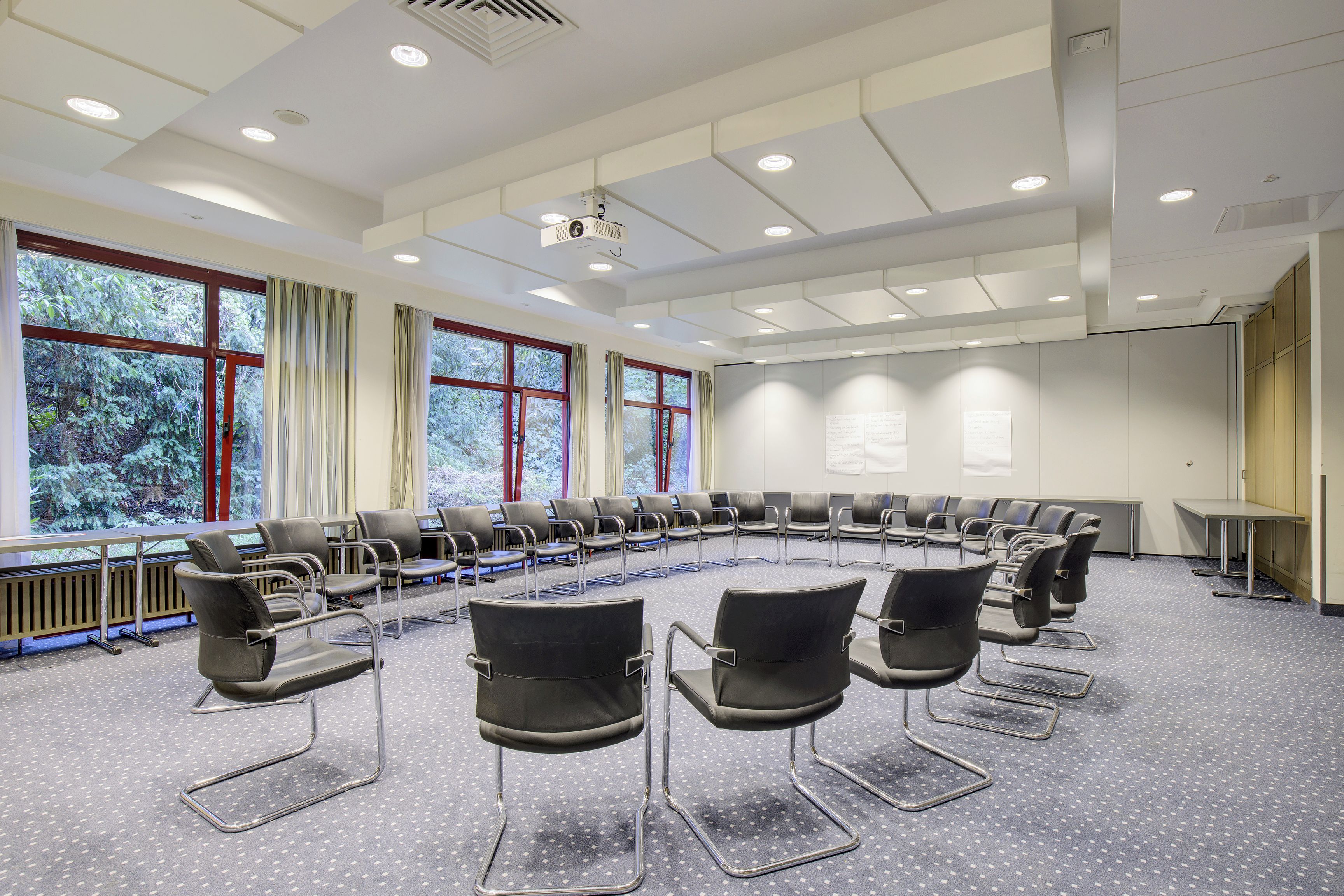 A conference room with a U-shaped seating arrangement, large windows with curtains, and a projector on the ceiling.