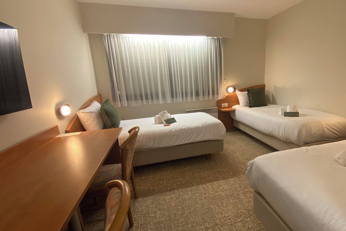A hotel room with two beds, a desk, and a chair. There are bedside tables with lamps and a window with curtains.