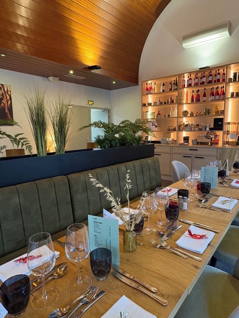 Um restaurante com uma longa mesa de madeira posta para refeição, um banco acolchoado verde e uma parede de garrafas de vinho.