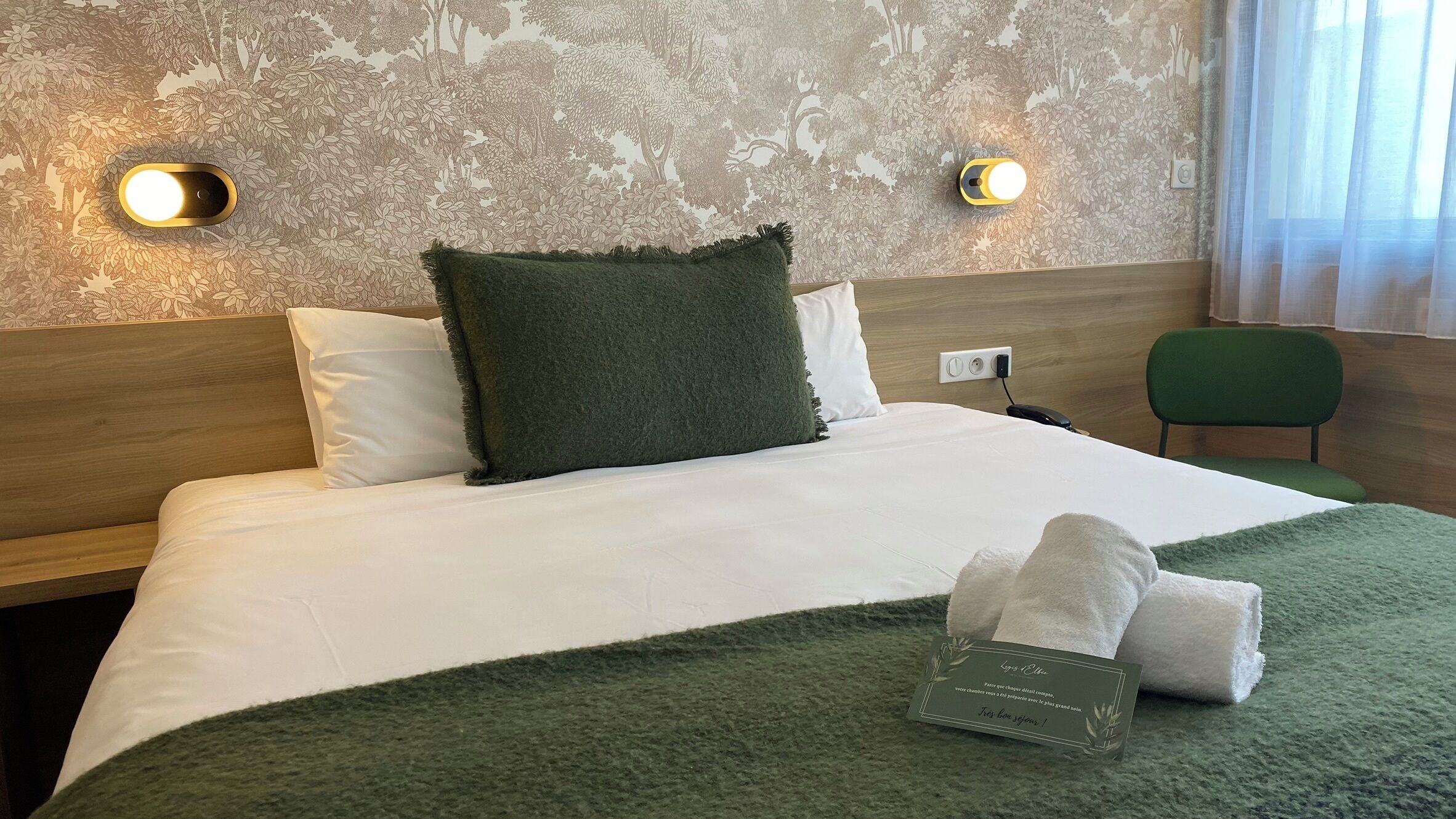 Una habitación de hotel con cama, dos lámparas de noche, una manta verde, una toalla y una tarjeta sobre la cama.