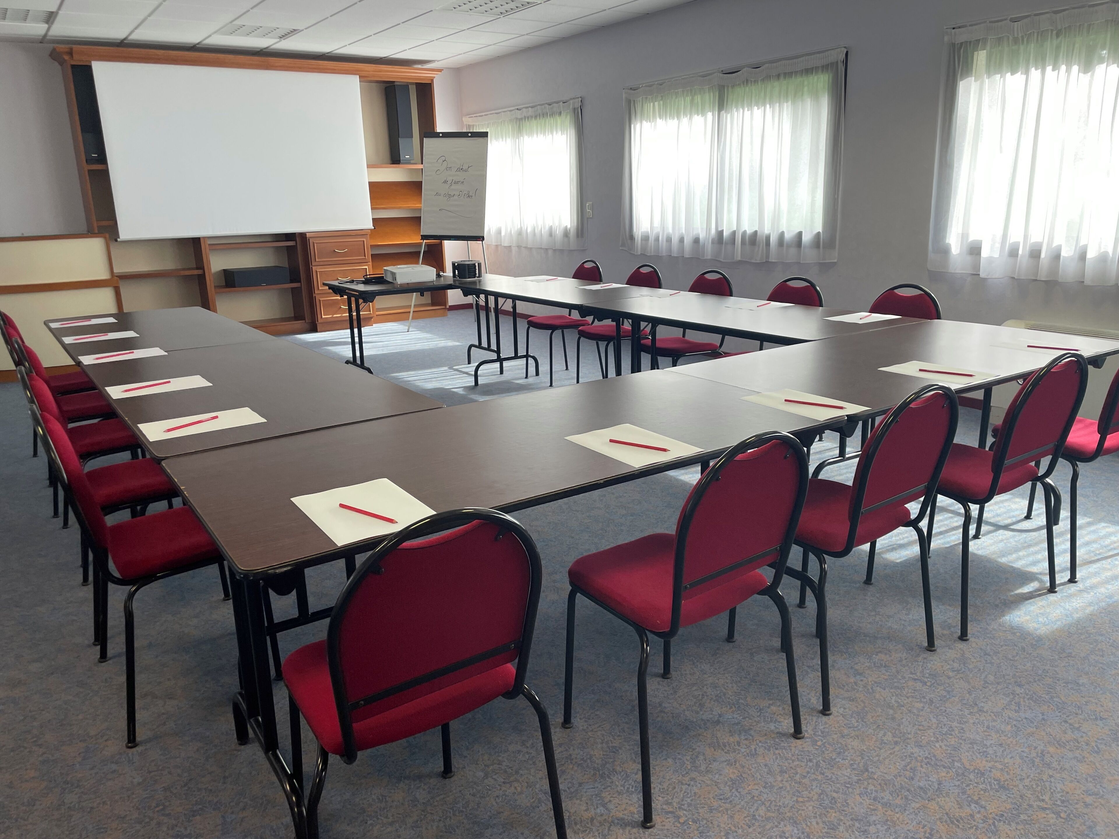 A conference room with long tables, red chairs, a whiteboard, and a projector screen.