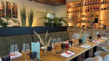 Un restaurante con una larga mesa de madera dispuesta para comer, una zona de bar con estantes llenos de botellas y plantas de interior.