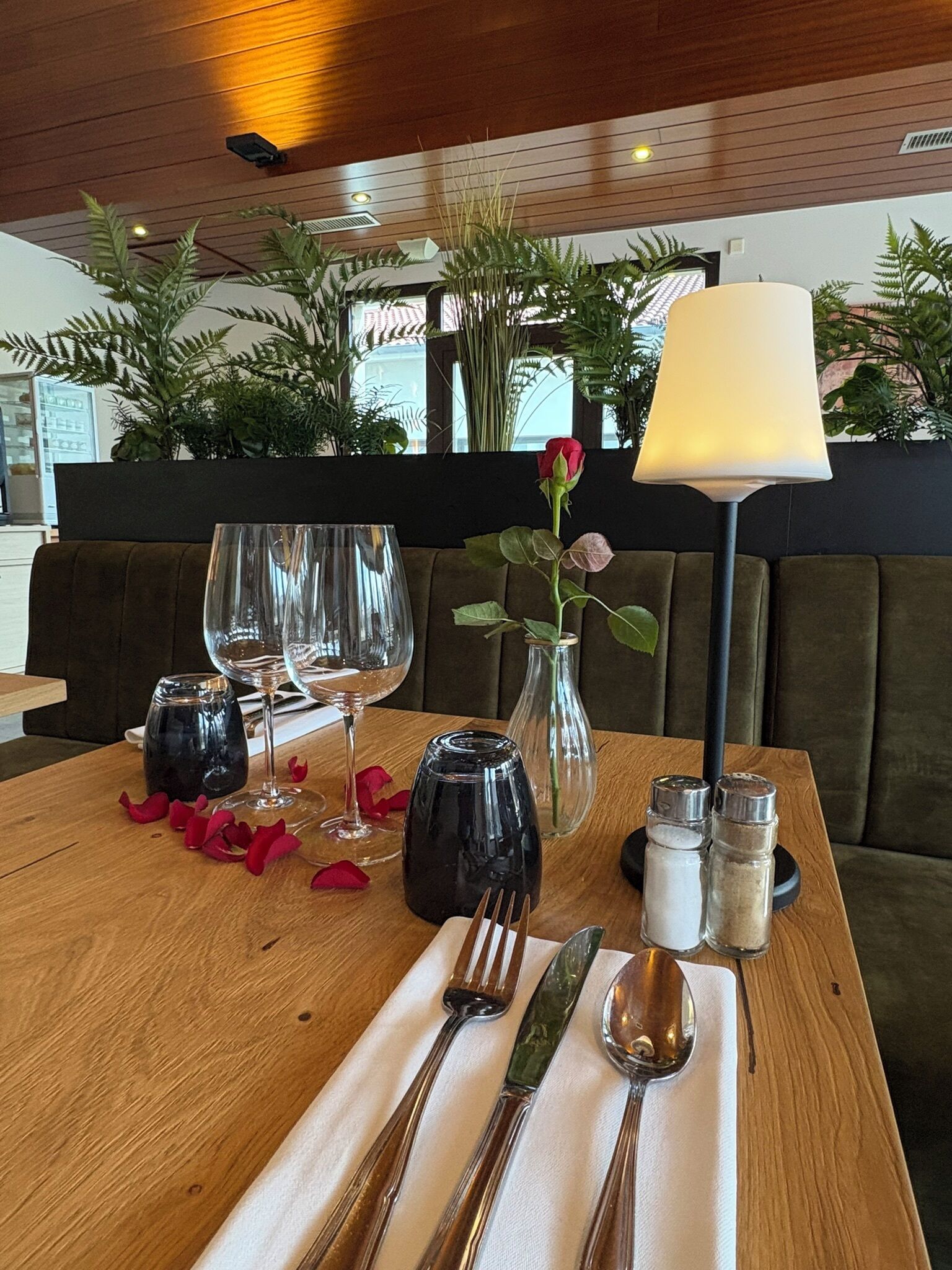 Una mesa de comedor dispuesta con copas de vino, un florero con una rosa, saleros y pimenteros, y una lámpara.