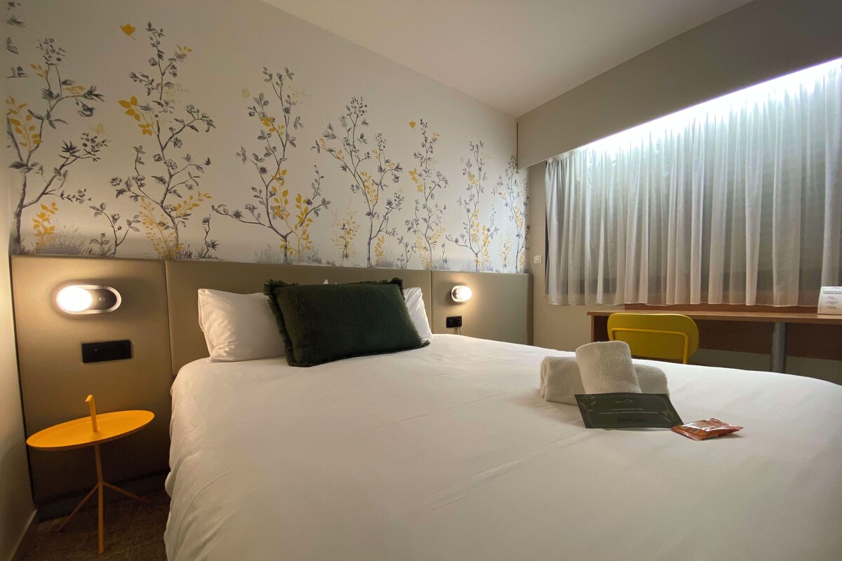 Quarto de hotel com uma cama grande, uma mesa redonda pequena, uma cadeira e uma janela com cortinas.