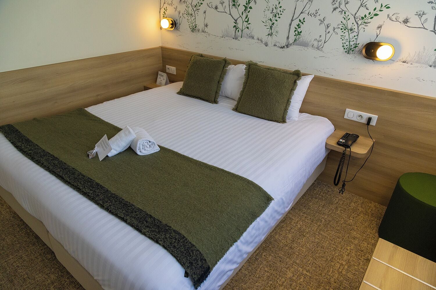 Una cama bien hecha con un plaid verde, un mando a distancia y un teléfono sobre la mesita de noche.