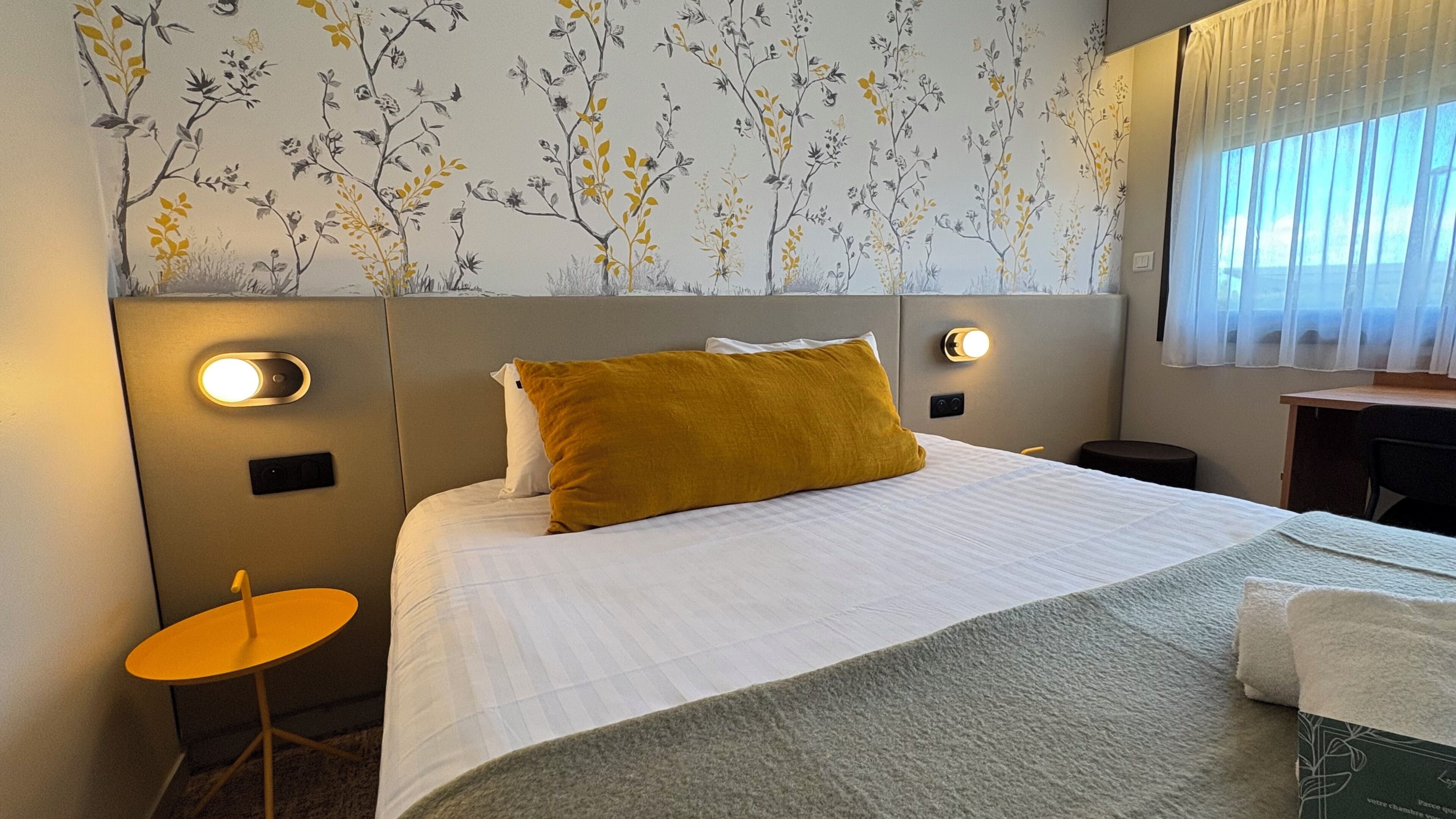 Ein Hotelzimmer mit Bett, einem kleinen runden Tisch, einem Schreibtisch und einem Fenster mit Vorhängen.