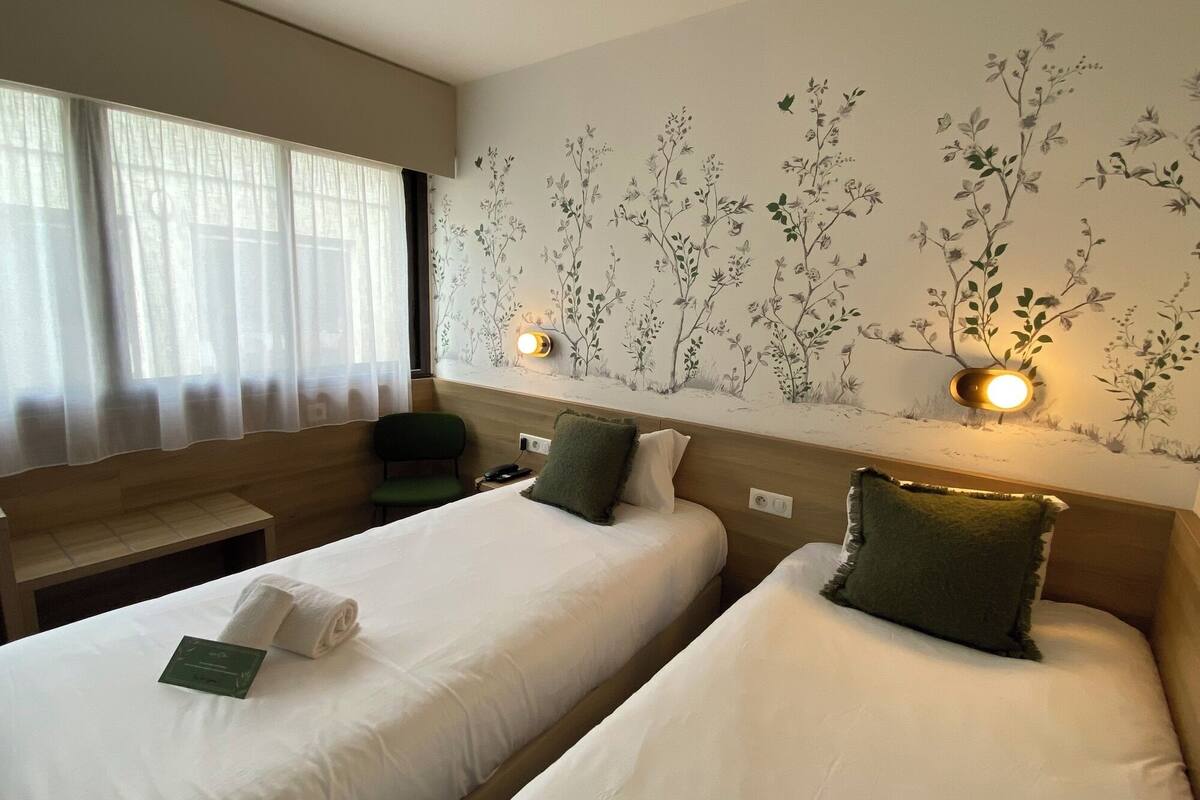 A hotel room with two single beds, a desk, a chair, and a wall with floral wallpaper.