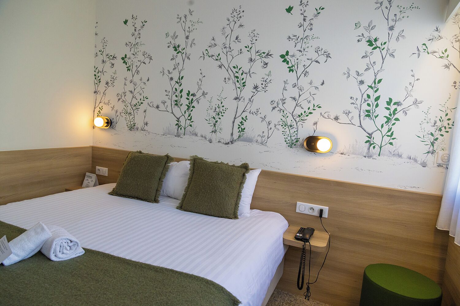 Una habitación de hotel con cama, dos almohadas, una mesita de noche con teléfono, una lámpara de pared y un cubo de basura verde.