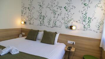 Una habitación de hotel con cama, dos almohadas, una mesita de noche con teléfono, una lámpara de pared y un cubo de basura verde.