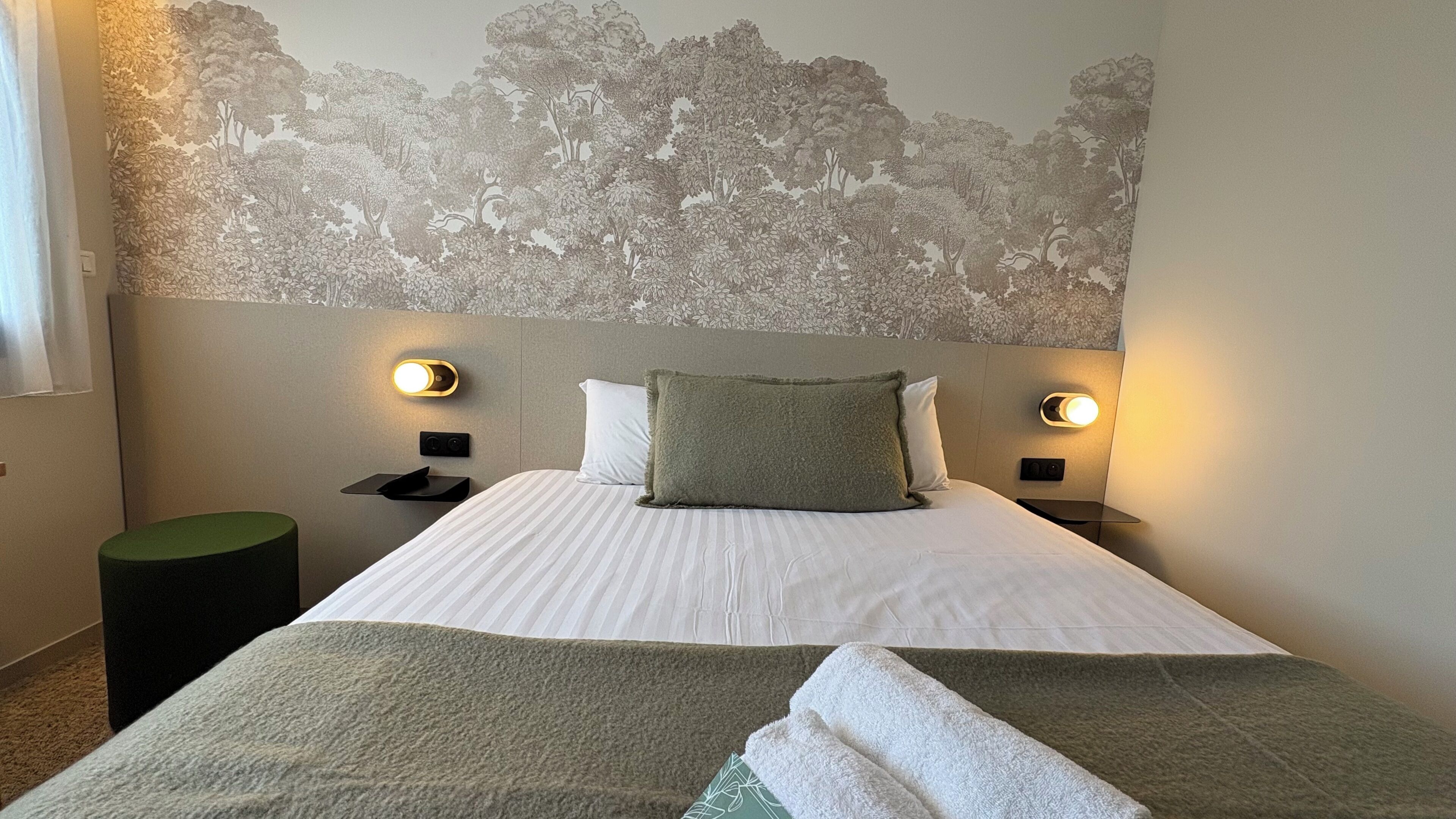 A hotel room with a bed, two bedside tables, a nightstand, a green ottoman, and a wall with a tree mural.