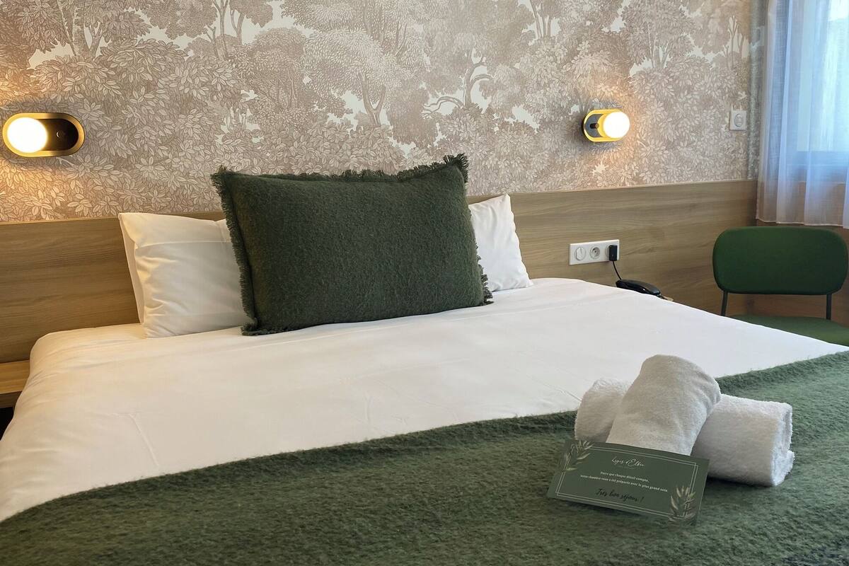 A neatly made bed with a green throw blanket and a set of towels. There are two wall-mounted lights and a power outlet on the wall.