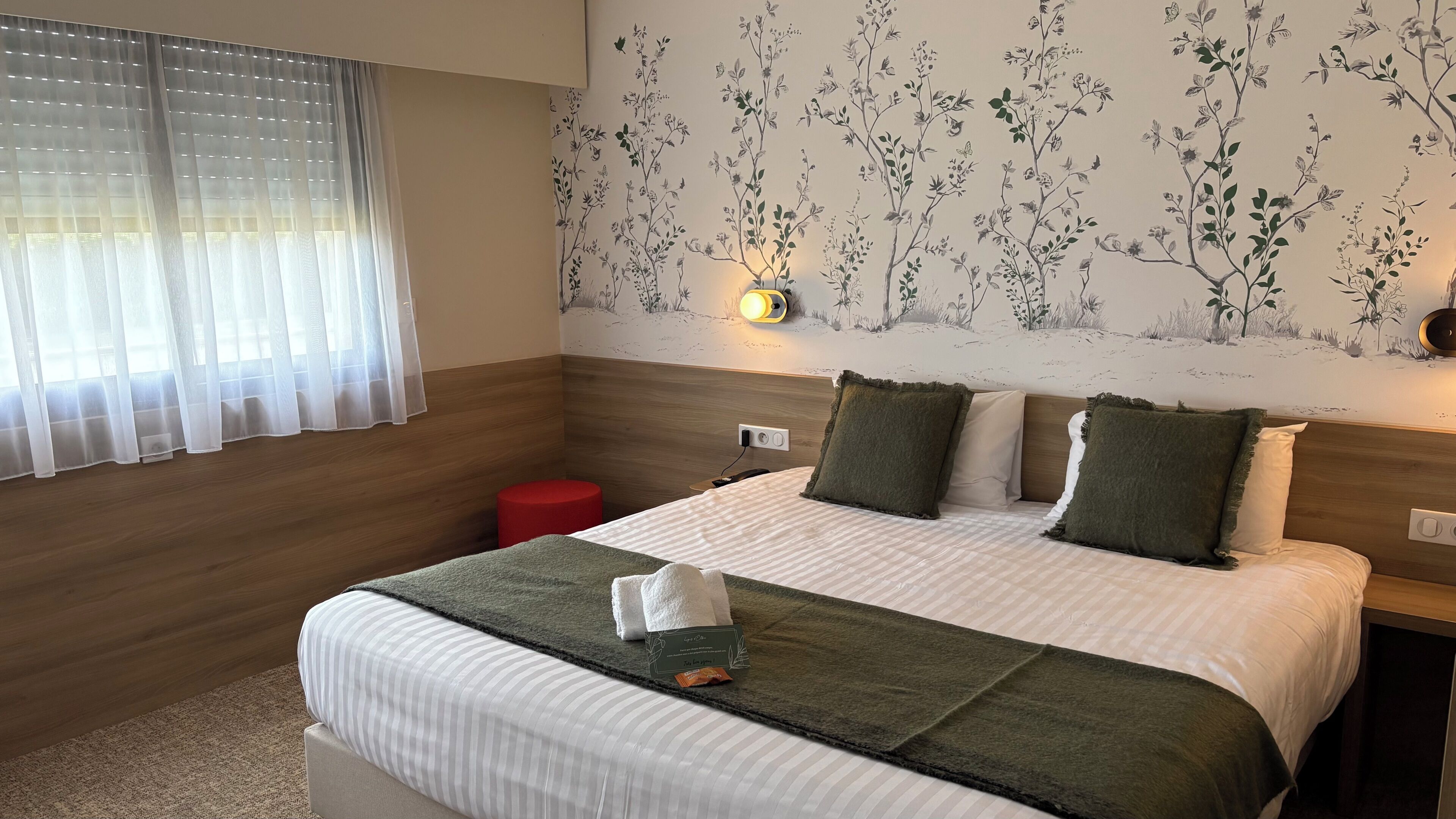 Habitación de hotel con cama, mesita de noche, un taburete rojo, una lámpara de pared y papel tapiz floral.