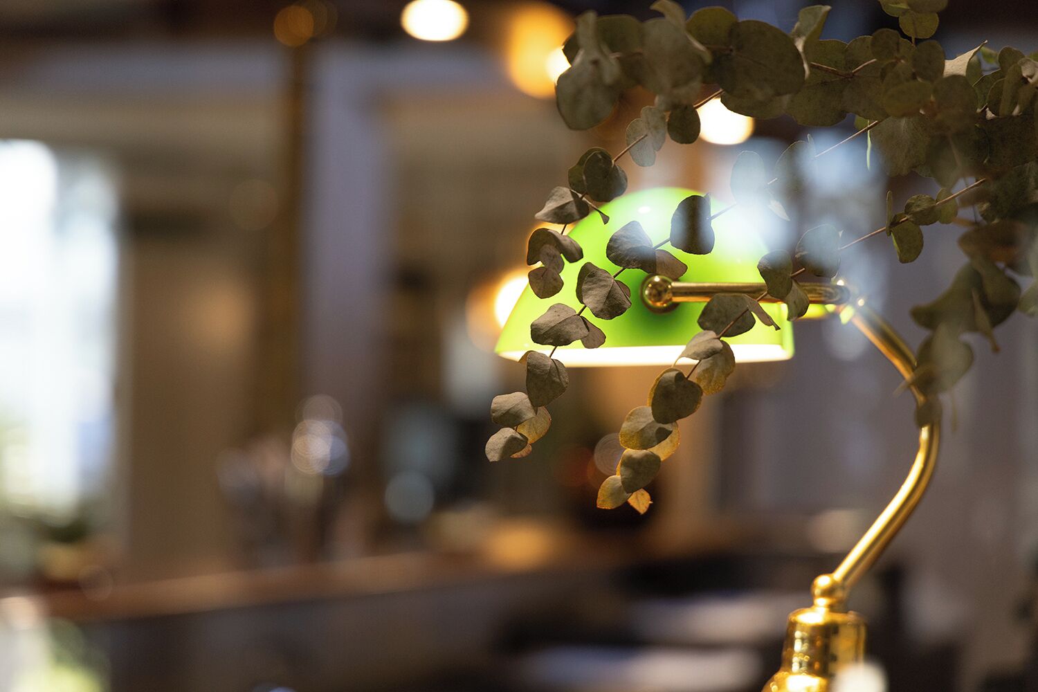 A lamp with a green shade and a gold base, with eucalyptus leaves in the foreground.