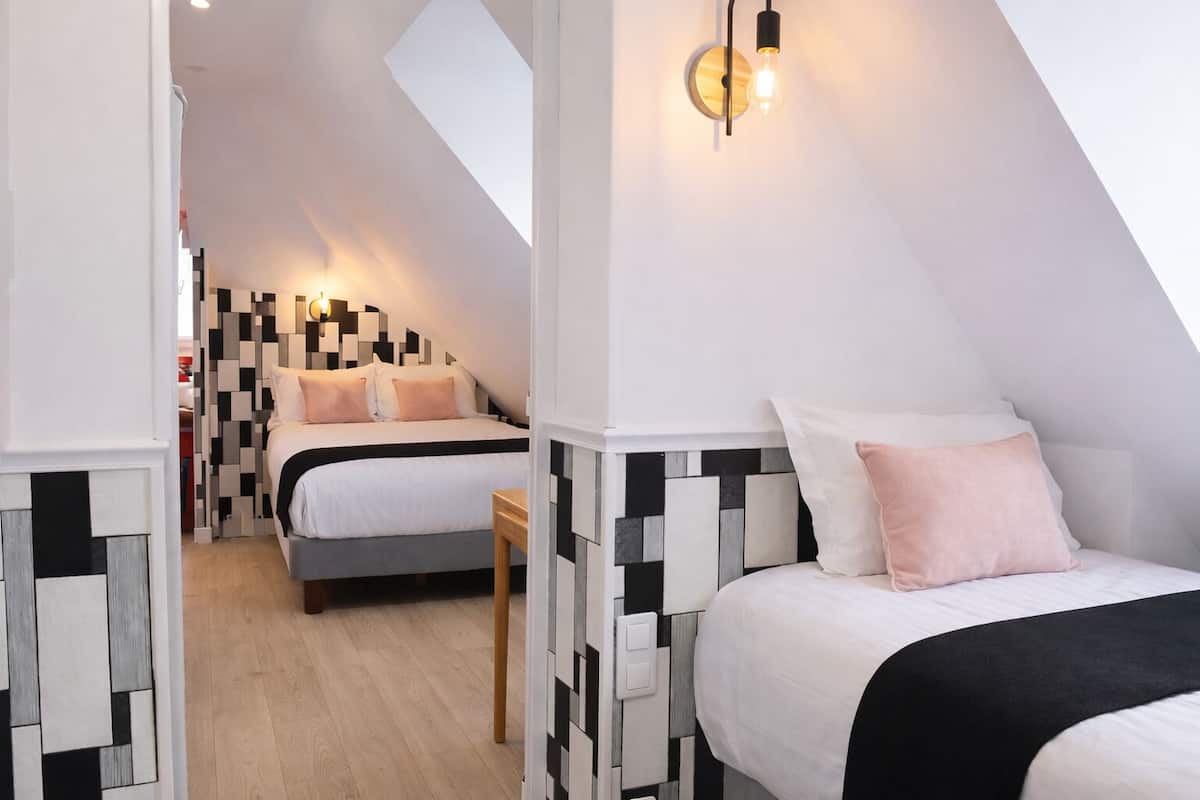 A modern hotel room with a sloped ceiling, two beds with black and white patterned headboards, a wooden floor, and a wall-mounted light fixture.