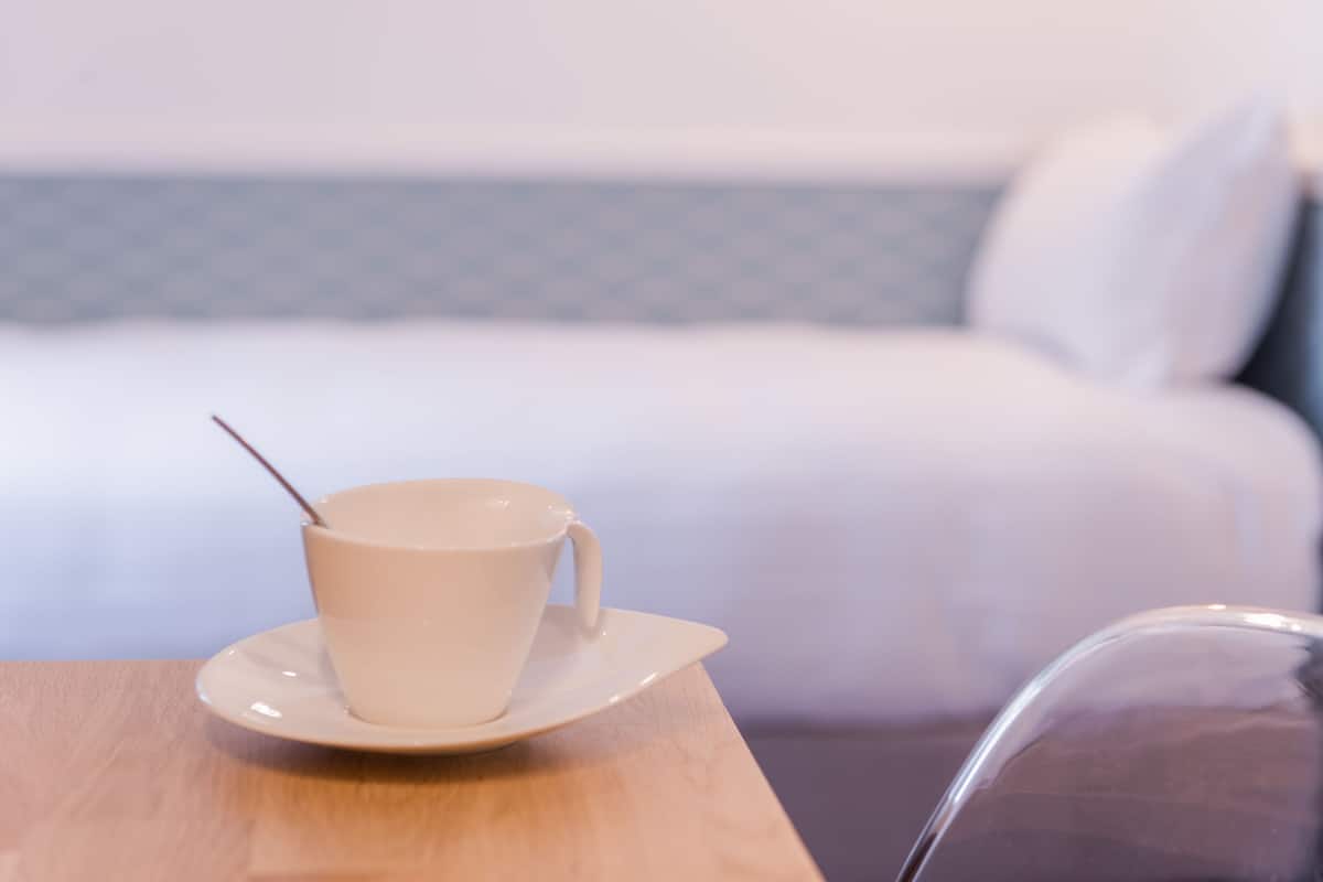 A hotel room with a bed in the background, a table with a cup and saucer, and a chair.