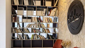 Un área de recepción de hotel con una estantería llena de libros, dos sillones rojos, una planta en maceta y un felpudo con el nombre del hotel.