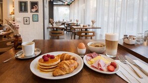 Un desayuno con panqueques, cruasanes, huevos revueltos, jamón, queso, fresas y bebidas sobre una mesa de madera en una cafetería con vistas al exterior.