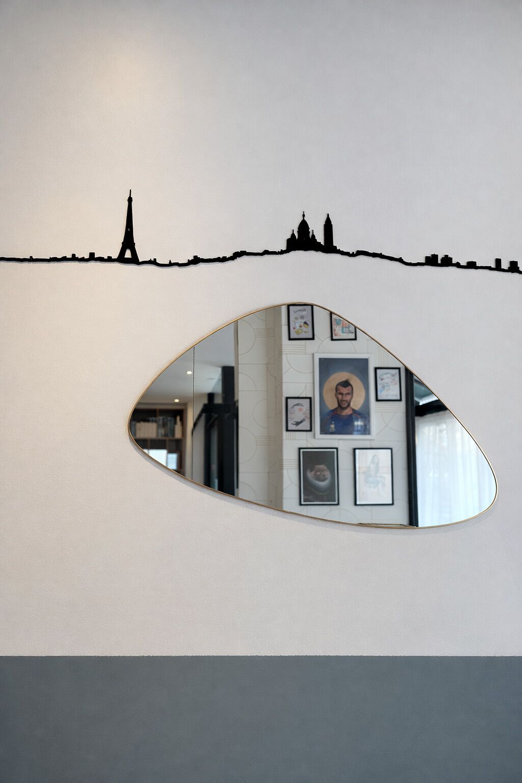 A wall with a silhouette of a city skyline, a mirror reflecting a framed portrait, and a partial view of a room with a bookshelf.