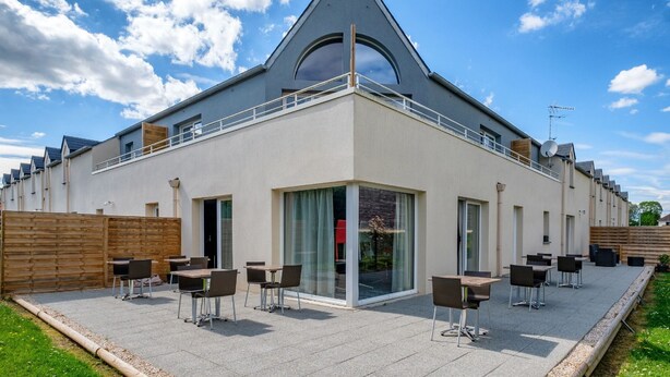 Il s’agit d’un bâtiment moderne doté d’un toit triangulaire et de grandes fenêtres. Il dispose d’un espace extérieur aménagé avec des tables et des chaises.