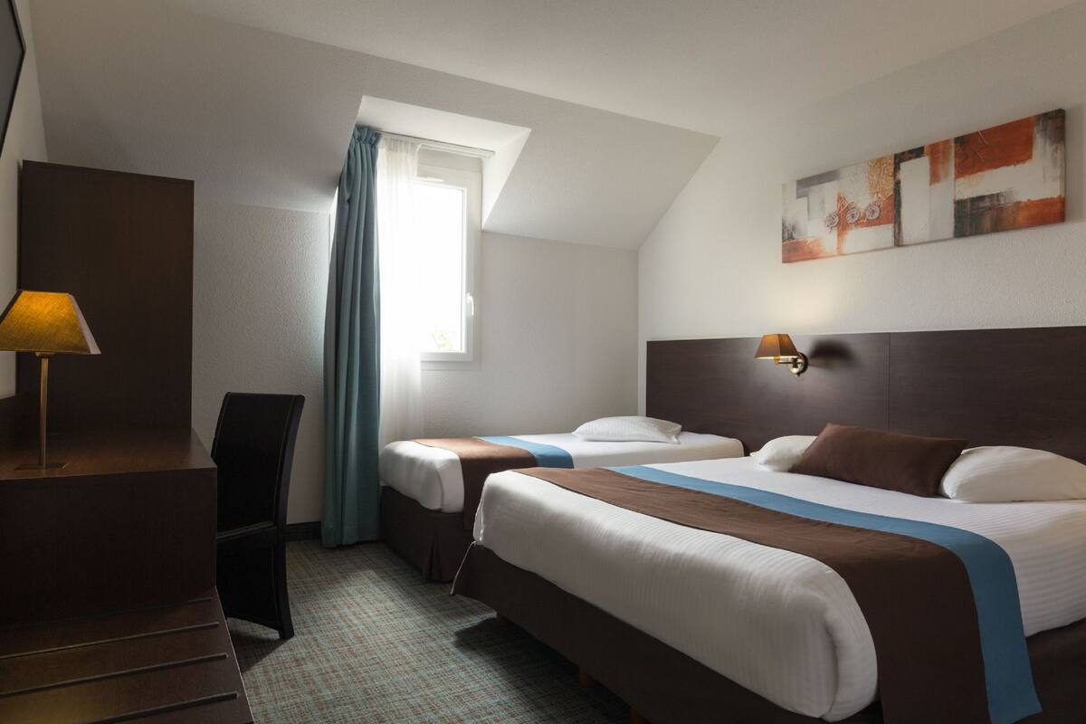 Habitación de hotel con dos camas, un escritorio, una lámpara y un ventanal con cortinas.