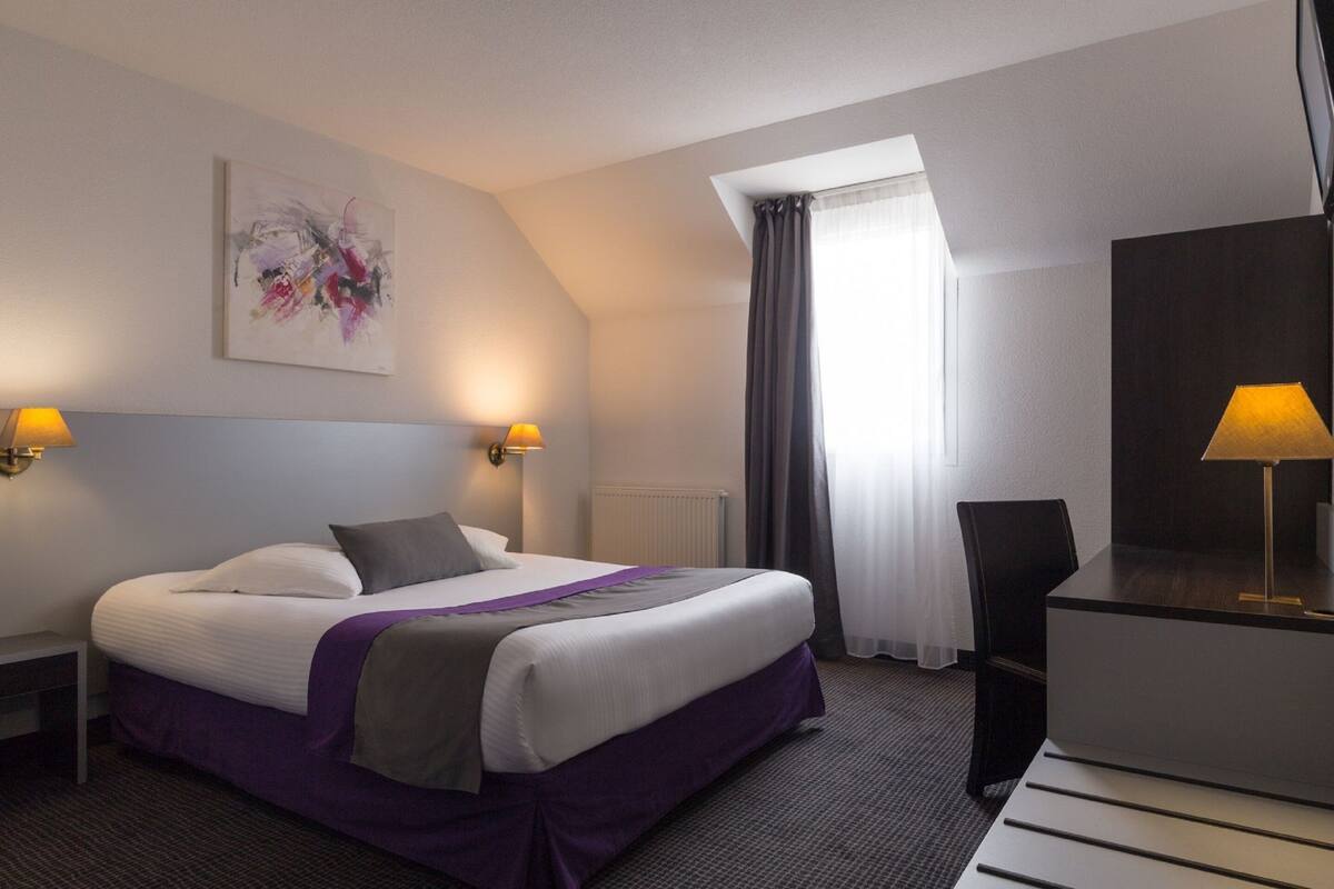 Habitación de hotel con cama, dos lámparas de noche, un escritorio con lámpara, una silla, un cuadro en la pared y una ventana con cortinas.