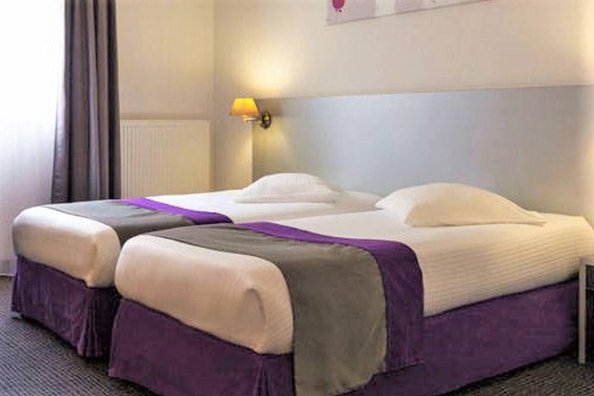 Habitación de hotel con dos camas, una mesita de noche, una lámpara y una ventana con cortinas.