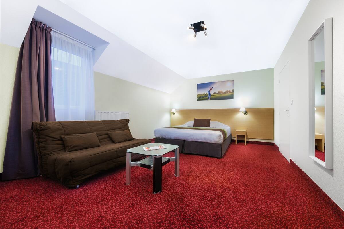 Habitación de hotel con alfombra roja, un sofá, una cama, una mesita y una lámpara en el techo.