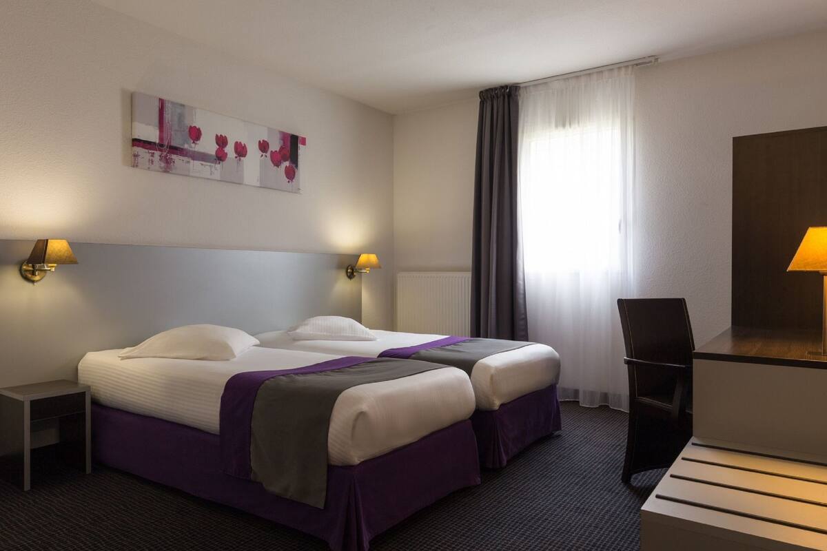 Habitación de hotel con dos camas, un escritorio, una silla, una lámpara y un cuadro en la pared.