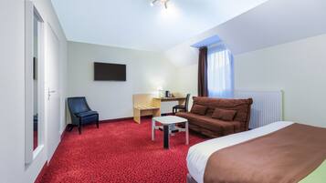 A hotel room with a red carpet, a bed, a desk, a chair, and a TV on the wall.