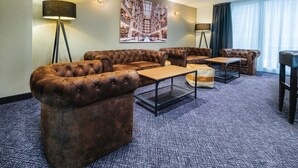 Un hall d’hôtel avec un grand canapé d’angle, une table basse et une petite table. On y trouve des lampadaires et un grand tableau accroché au mur.