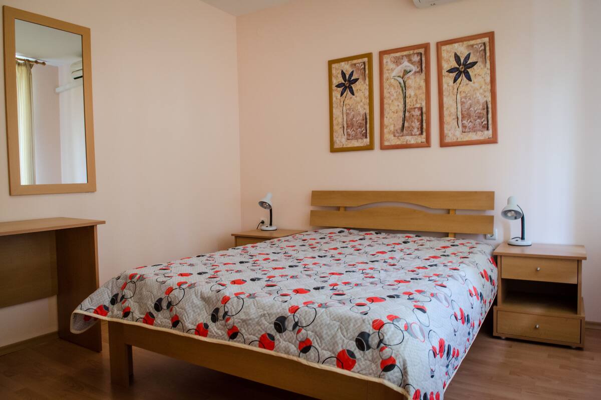 A bedroom with a bed, bedside tables, a desk, and a mirror. There are three framed pictures on the wall.