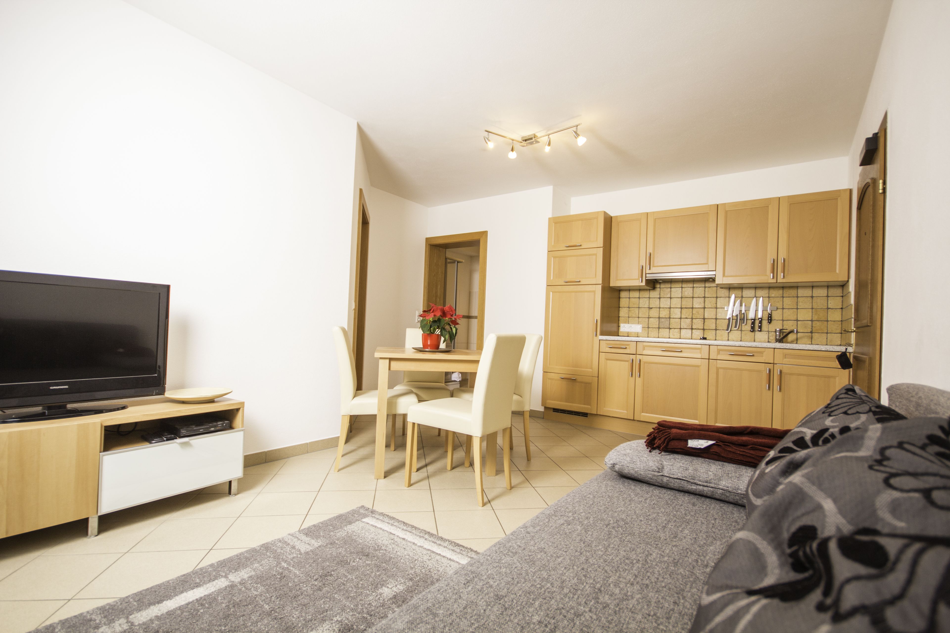 Un espacio habitable compacto con televisión, mesa de comedor, sillas, sofá y zona de cocina con armarios de madera y salpicadero de azulejos.
