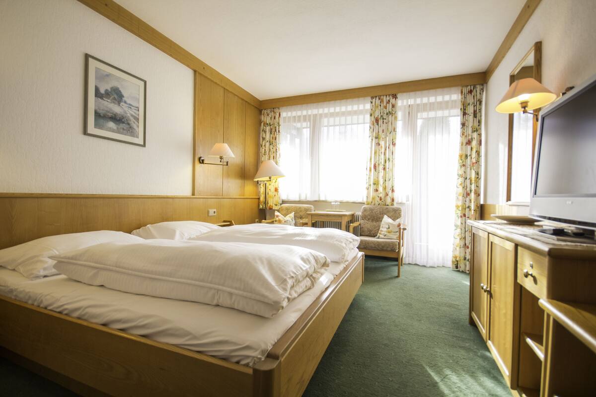 Quarto de hotel com cama de madeira, escrivaninha, cadeira, televisão e janela com cortinas florais.