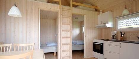 A compact wooden cabin with a dining area, a loft bedroom with a ladder, a kitchenette, and a window with blinds.