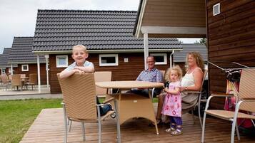 Eine Familie auf einer Holzterrasse mit Gartenmöbeln, einem überdachten Bereich und einem Gebäude mit Ziegeldach im Hintergrund.