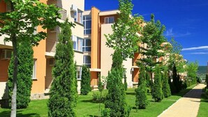 Un immeuble résidentiel à plusieurs étages, doté de balcons, entouré d’un espace vert bien entretenu et d’un chemin piétonnier dallé.