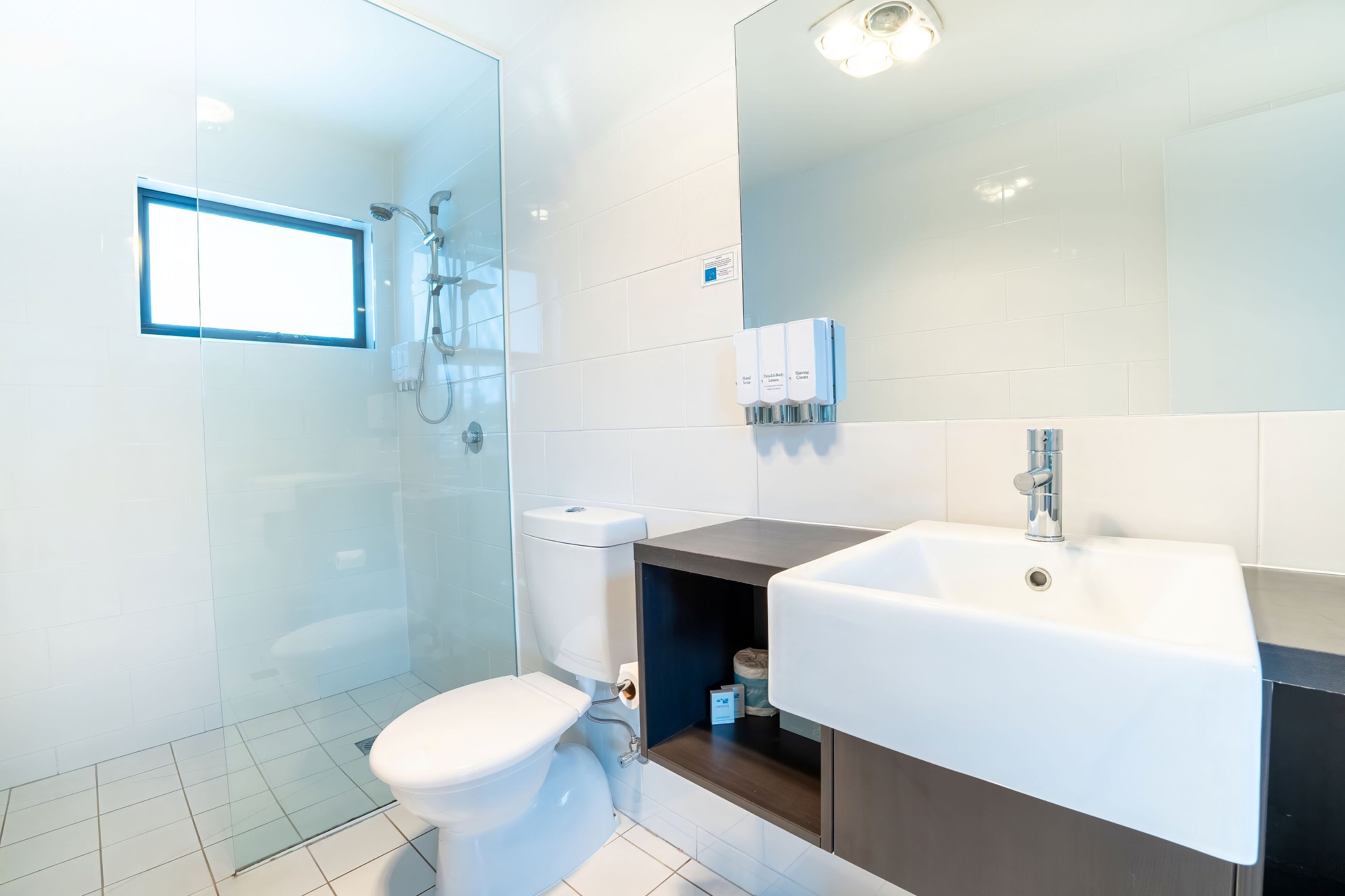 Un baño moderno con cabina de ducha de vidrio, inodoro blanco, lavamanos con espejo y un estante pequeño con artículos de tocador.