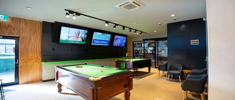 A pool table with two screens displaying sports content, a bar area, and seating.