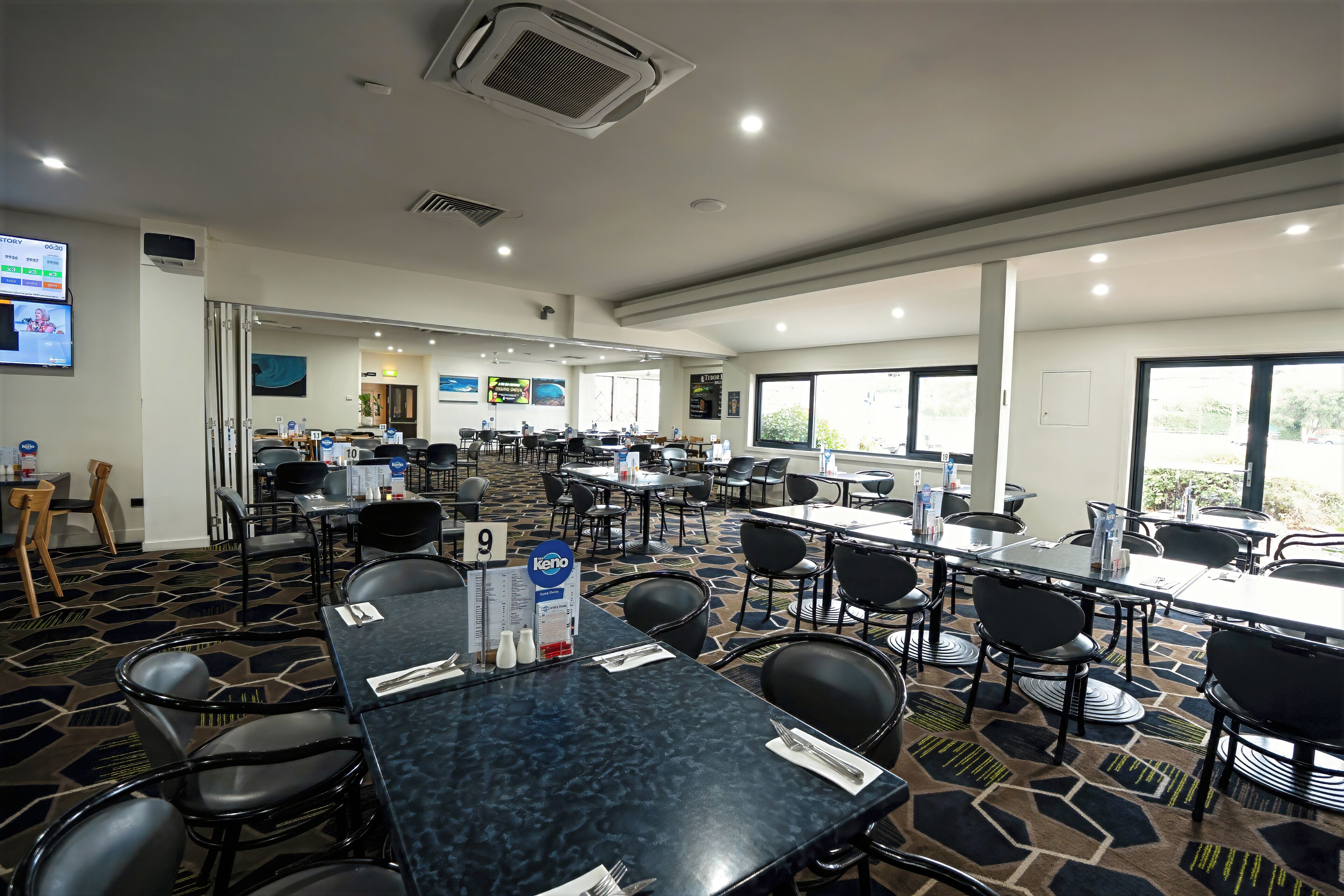 Een restaurantgedeelte in een hotel met meerdere tafels en stoelen, een groot raam en een scherm dat informatie toont.