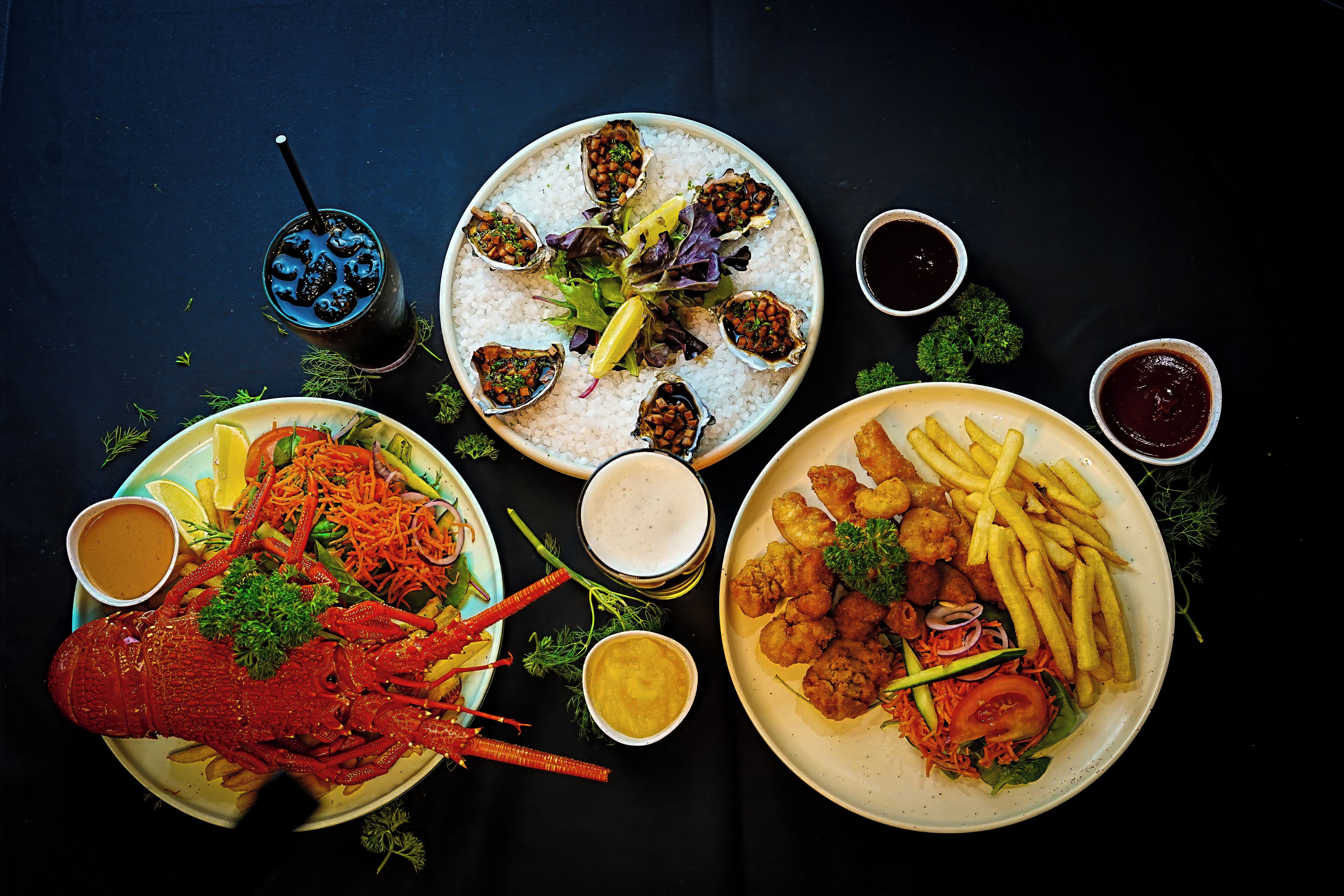 A table with seafood dishes, including lobster, oysters, and fried items, accompanied by various sauces and a drink.