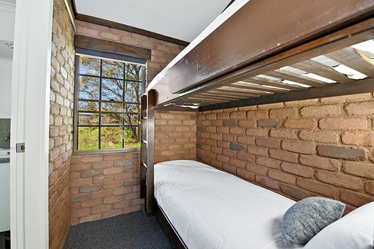 A small, single-occupancy room with a bunk bed, a window with a view of trees, and a brick wall.