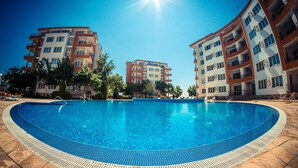 A swimming pool surrounded by multi-story buildings with balconies, a clear sky, and a sun shining overhead. - Riviera Fort Beach (Ravda)