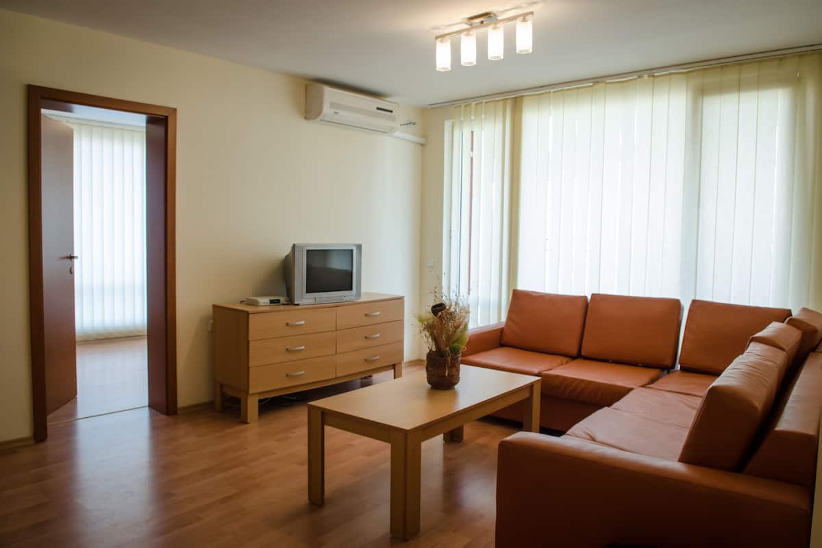 A living room with a sofa, a coffee table, a TV on a wooden cabinet, and a door leading to another room.