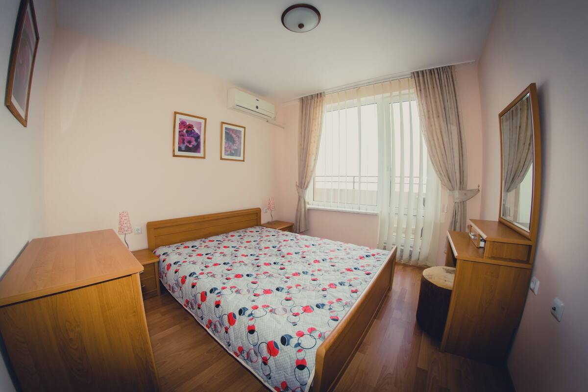 Un dormitorio con cama de madera, una mesita de noche, un tocador, un espejo y una ventana con cortinas.
