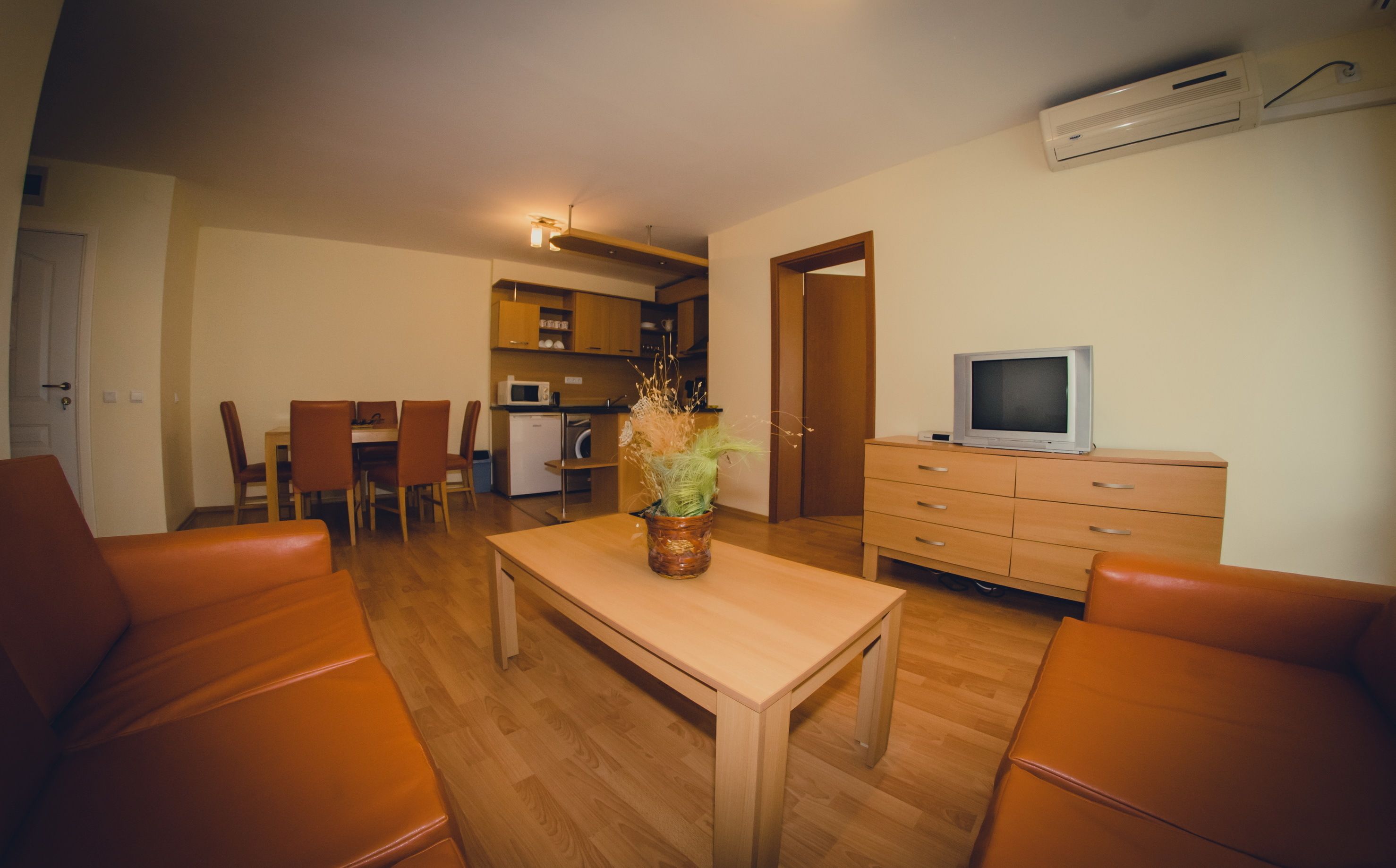 Een woonkamer met een houten tafel, oranje stoelen, een televisie op een dressoir en een keuken op de achtergrond.