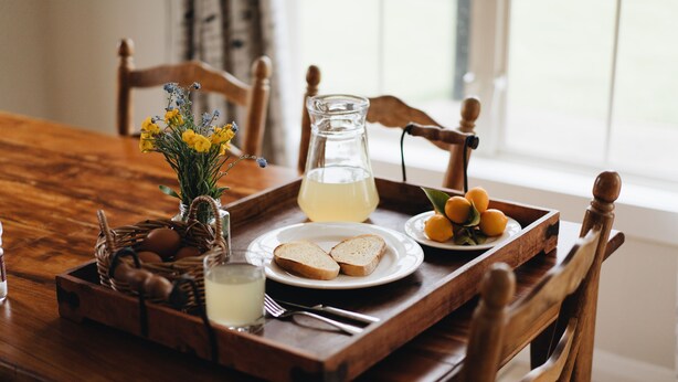 A wooden dining table with a tray containing bread, a pitcher of lemonade, a basket of eggs, and a vase of flowers.