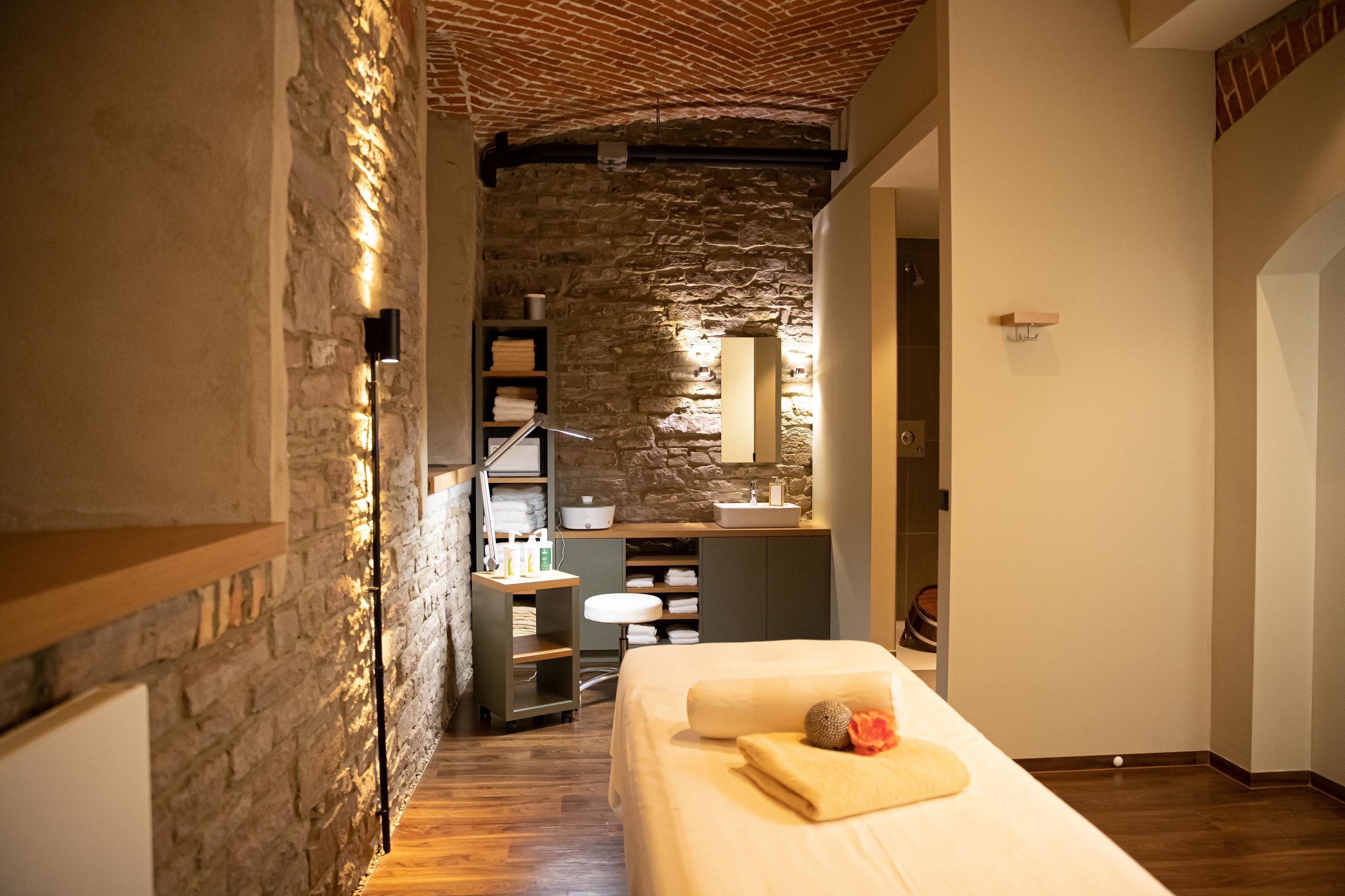 A cozy room with a stone wall, wooden flooring, a bed with a towel and fruit, a small bathroom with a sink and shelves.